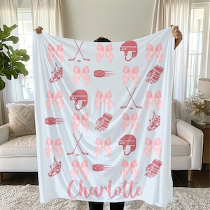 May include: White blanket with a pink hockey theme. The blanket features pink hockey equipment, bows, and the name "Charlotte" in a script font. The design includes hockey helmets, skates, pucks, and gloves.
