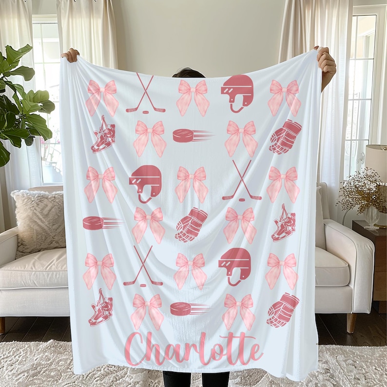 May include: White blanket with a pink hockey theme. The blanket features pink hockey equipment, bows, and the name "Charlotte" in a script font. The design includes hockey helmets, skates, pucks, and gloves.
