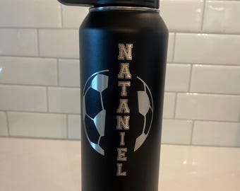 May include: Black insulated water bottle with a black handle and lid. The bottle features the name "Nataniel" in vertical silver lettering, with a soccer ball design. The bottle is designed to keep drinks cold.