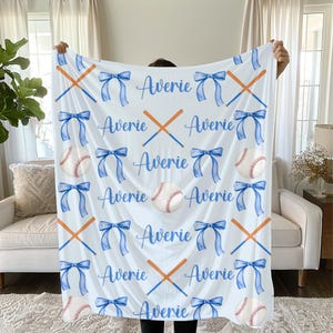 Puede incluir: Manta blanca con temática de béisbol con lazos azules, pelotas de béisbol y bates cruzados. El nombre "Averie" está escrito en azul. La manta está sostenida frente a una persona.
