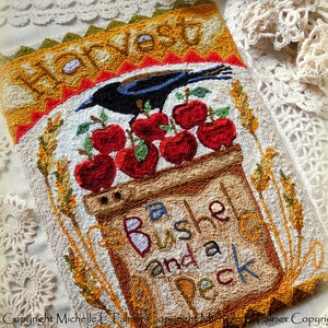 Puede incluir: Un tapiz de punto de aguja con un tema de cosecha. Presenta un cuervo posado en una cesta de bushel llena de manzanas rojas. El texto "Harvest a Bushel and a Peck" está cosido en hilo marrón y blanco.