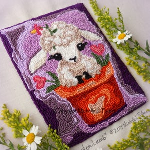 May include: A needle felted lamb in a flower pot with pink and green flowers. The lamb is white with black eyes and a brown nose. The pot is orange with a heart on the side. The background is purple.