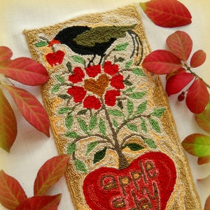 Puede incluir: Un diseño de punto de aguja de una manzana con las palabras "apple a day" en su interior. Un pájaro negro se posa en una rama con flores rojas y hojas verdes. El diseño está rodeado por un borde de color crema. El texto "Copyright Michelle L. Palmer" y "Apple a day ©2017 Michelle L. Palmer" está en la parte inferior del diseño.