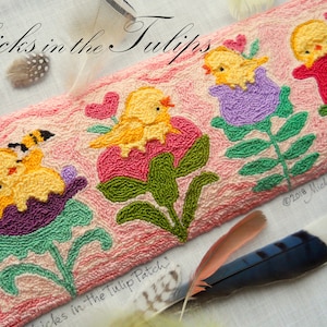 Peut inclure: Un coussin en point d'aiguille rose et blanc avec un motif brodé de poussins jaunes dans des tulipes violettes et roses. Le coussin porte le texte "Chicks in the Tulips" et "Chicks in the Tulip Patch" brodé dessus.