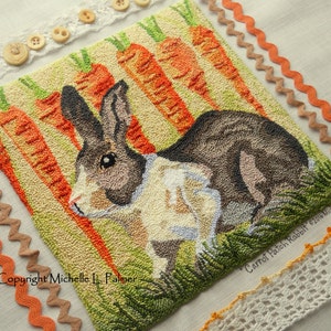 May include: A needlepoint embroidery of a brown and white rabbit sitting in a field of green grass and orange carrots. The text "Carrot Patch Rabbit" is in the lower right corner of the image. The text "Copyright Michelle L. Palmer" is in the lower left corner of the image.