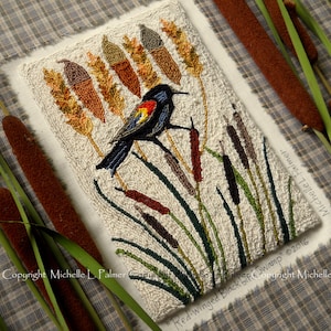 May include: Needlepoint artwork depicting a red-winged blackbird perched on cattails. The bird is black with red patches on its wings. The cattails are brown and green. The background is white.