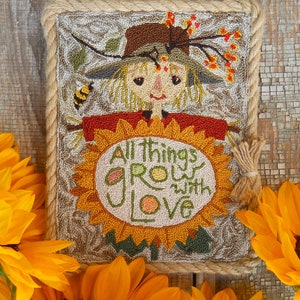 May include: A framed, textured artwork featuring a smiling scarecrow with a sunflower. The scarecrow wears a brown hat and red shirt, with the text "All things grow with love" on the sunflower. The artwork is framed with rope and surrounded by sunflowers.