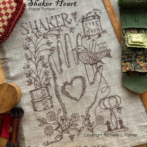 May include: A linen fabric with a brown embroidered design featuring the words "Shaker Heart" and "SHAKER". The design includes a bird, a house, a heart, and floral elements. The pattern measures 11.5" x 15.5".