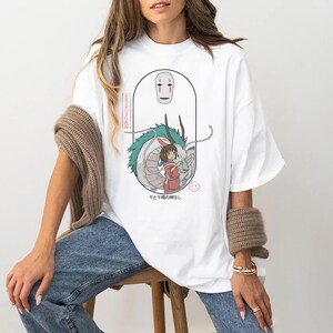 Puede incluir: Camiseta blanca con un estampado gr&aacute;fico de un personaje de la pel&iacute;cula de animaci&oacute;n El viaje de Chihiro. El gr&aacute;fico muestra un drag&oacute;n, una ni&ntilde;a y una m&aacute;scara. El texto en la camiseta dice "That's the spirit!"