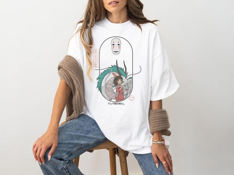 Puede incluir: Camiseta blanca con un estampado gr&aacute;fico de un personaje de la pel&iacute;cula de animaci&oacute;n El viaje de Chihiro. El gr&aacute;fico muestra un drag&oacute;n, una ni&ntilde;a y una m&aacute;scara. El texto en la camiseta dice "That's the spirit!"