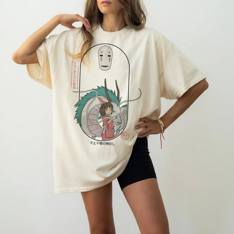 Puede incluir: Una camiseta de color crema con un estampado gr&aacute;fico de un drag&oacute;n, una ni&ntilde;a y una figura enmascarada. El texto en la camiseta dice "スタジオジブリ" y "千と千尋の神隠し".