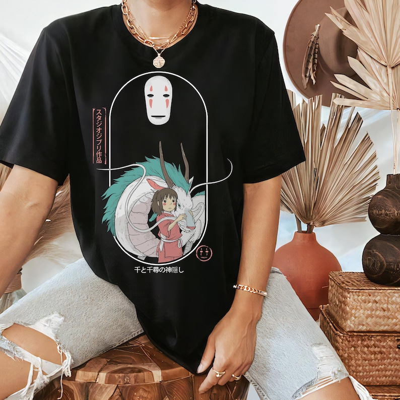 May include: Black t-shirt featuring Spirited Away artwork.  The design includes Chihiro, No-Face, and Haku the dragon. Japanese text is present on the shirt.  The graphic is a stylized illustration in muted colors against the black fabric.