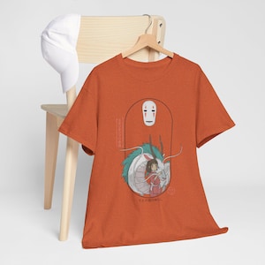 Puede incluir: Camiseta naranja con un estampado gr&aacute;fico de un drag&oacute;n blanco y una ni&ntilde;a con un vestido rojo. El drag&oacute;n tiene escamas verdes y azules. El texto en la camiseta dice "No Face" y "Con el viento del d&iacute;a".