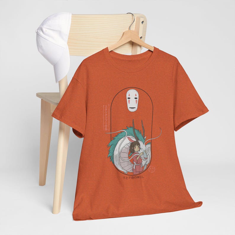 Puede incluir: Camiseta naranja con un estampado gr&aacute;fico de un drag&oacute;n blanco y una ni&ntilde;a con un vestido rojo. El drag&oacute;n tiene escamas verdes y azules. El texto en la camiseta dice "No Face" y "Con el viento del d&iacute;a".