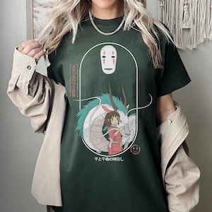 Puede incluir: Una camiseta verde oscuro con un estampado gr&aacute;fico de un drag&oacute;n blanco, una ni&ntilde;a y una m&aacute;scara de Sin Cara de la pel&iacute;cula El viaje de Chihiro. El texto japon&eacute;s en la camiseta dice "Sen to Chihiro no Kamikakushi", que se traduce como "El viaje de Chihiro".