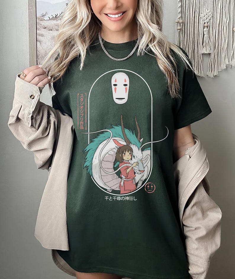 Puede incluir: Una camiseta verde oscuro con un estampado gr&aacute;fico de un drag&oacute;n blanco, una ni&ntilde;a y una m&aacute;scara de Sin Cara de la pel&iacute;cula El viaje de Chihiro. El texto japon&eacute;s en la camiseta dice "Sen to Chihiro no Kamikakushi", que se traduce como "El viaje de Chihiro".