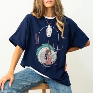 Puede incluir: Una camiseta azul marino con un estampado gr&aacute;fico de un drag&oacute;n blanco, una ni&ntilde;a y una m&aacute;scara de No Face. El texto en la camiseta dice "スタジオジブリ作品 千と千尋の神隠し".