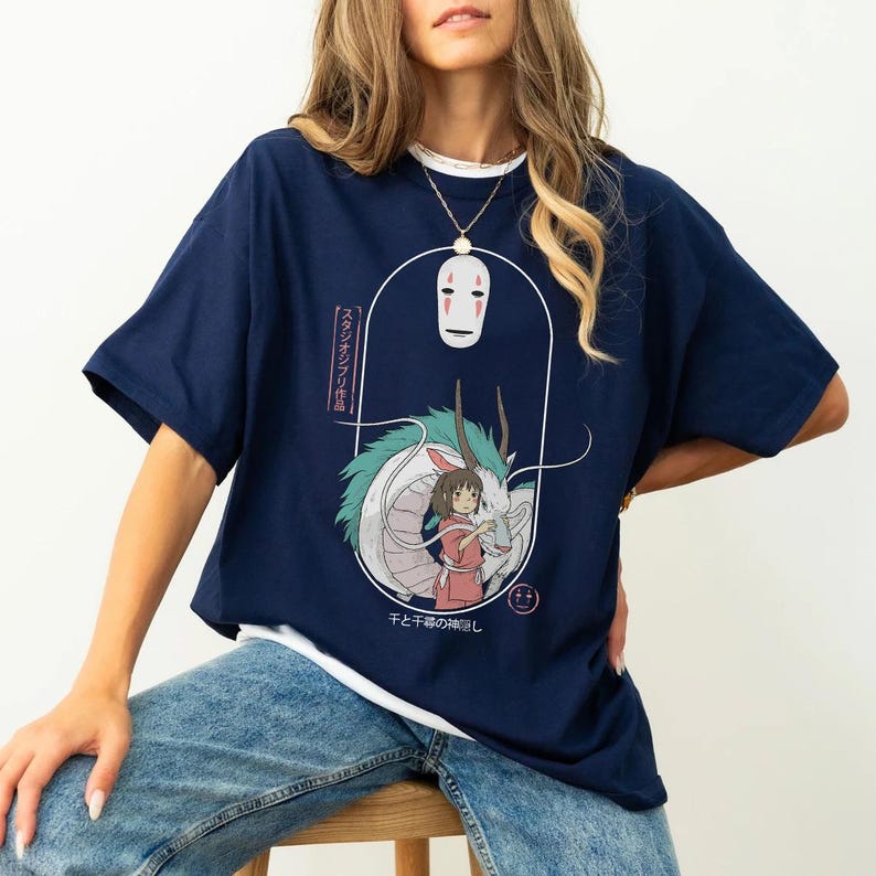 Puede incluir: Una camiseta azul marino con un estampado gr&aacute;fico de un drag&oacute;n blanco, una ni&ntilde;a y una m&aacute;scara de No Face. El texto en la camiseta dice "スタジオジブリ作品 千と千尋の神隠し".