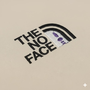 Op de afbeelding: Beige stof met de zwarte borduursels "THE NO FACE". Het ontwerp bevat een gestileerd gezicht met een zwarte boogvormige omtrek en paarse accenten, dat doet denken aan een stripfiguur.