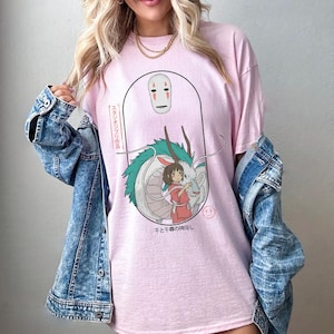 Puede incluir: Una camiseta rosa con un estampado gr&aacute;fico de una m&aacute;scara de esp&iacute;ritu blanco, un drag&oacute;n verde y una ni&ntilde;a. El texto japon&eacute;s en la camiseta dice "Sen to Chihiro no Kamikakushi", que se traduce como "El viaje de Chihiro".