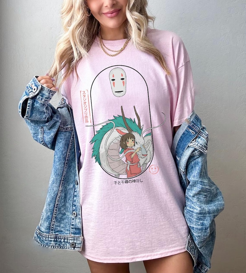 Puede incluir: Una camiseta rosa con un estampado gr&aacute;fico de una m&aacute;scara de esp&iacute;ritu blanco, un drag&oacute;n verde y una ni&ntilde;a. El texto japon&eacute;s en la camiseta dice "Sen to Chihiro no Kamikakushi", que se traduce como "El viaje de Chihiro".