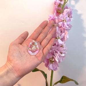 May include: A clear, iridescent teardrop-shaped pendant with an oval design in the center. The pendant is held in a hand, with a backdrop of pink flowers. The pendant reflects multiple colors, including pink, purple, and yellow.
