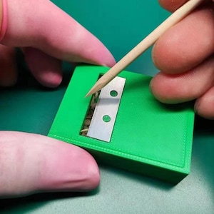 Peut inclure: Un objet rectangulaire vert vif avec une lame argentée et deux trous circulaires. Un cure-dent en bois est utilisé pour manipuler le contenu à l'intérieur. L'objet est sur une surface verte.