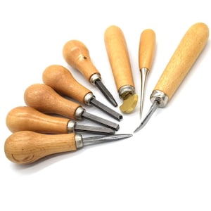 Può includere: Una collezione di strumenti per la lavorazione della pelle con manici in legno e lame in metallo. Gli strumenti includono varie forme, come una lama curva, un lesina appuntita e uno strumento a bordo arrotondato. Gli strumenti sono disposti su una superficie bianca.