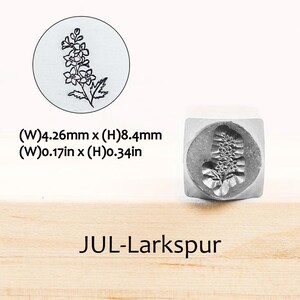 Pode incluir: Um carimbo de metal com um desenho floral de espargos. O carimbo tem 4,26 mm de largura e 8,4 mm de altura. O texto "JUL-Larkspur" est&aacute; impresso abaixo.