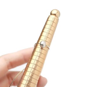 May include: A gold-colored ring sizer with numbers and a ring with a silver band and a pearl-shaped bead. The ring is on the sizer, which is held by a hand. The sizer has numbers from 1 to 18.