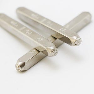 Könnte beinhalten: Nahaufnahme von drei Metallstempeln mit astrologischen Symbolen und Zahlen. Die silberfarbenen Stempel haben einen rechteckigen Körper und ein Muster am Stempelende. Die Stempel sind überlappend vor einem weißen Hintergrund angeordnet.