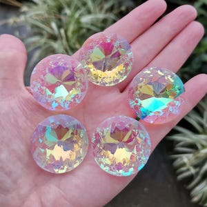 Puede incluir: Cinco piedras de cristal redondas e iridiscentes con un diseño facetado. Los cristales muestran un arcoíris de colores, incluyendo rosa, amarillo y azul. Las piedras se sostienen en una mano, con un fondo verde borroso.