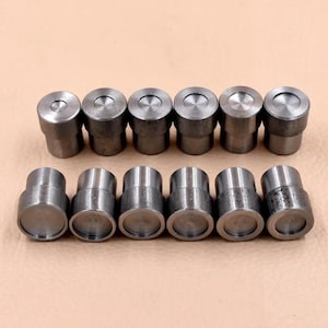 May include: A collection of twelve cylindrical metal objects, arranged in two rows of six. Each object has a flat top and a slightly textured, metallic finish. The items are evenly spaced against a neutral background.