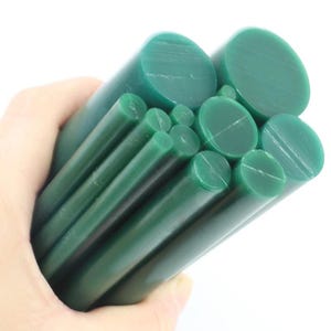 Puede incluir: Un conjunto de barras de cera verde oscuro de varios diámetros. Las barras cilíndricas se sostienen en una mano sobre un fondo blanco. Los extremos de las barras son visibles, mostrando una textura lisa y sólida. Las barras de cera son de color verde oscuro.