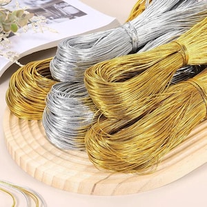 Puede incluir: Primer plano de cordones metálicos dorados y plateados, enrollados y agrupados en una bandeja de madera. Los cordones dorados tienen un tono cálido y rico, mientras que los cordones plateados ofrecen un contraste frío. Ideal para manualidades.