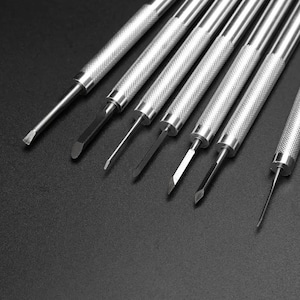 以下が含まれることがあります： テクスチャ加工されたハンドルとさまざまなブレード形状を備えた、7つの銀色の金属製ツールのセット。ツールは暗い背景に対して斜めに配置されており、平らな、角度の付いた、尖ったデザインなど、さまざまな先端を示しています。これらのツールは、詳細な工芸品または修理作業に使用される可能性があります。