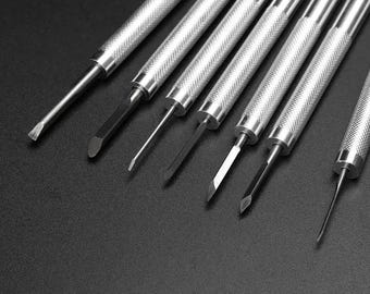 May include: A set of seven silver metal tools with textured handles and various blade shapes. The tools are arranged diagonally against a dark background, showcasing their different tips, including flat, angled, and pointed designs. These tools are likely for detailed craft or repair work.