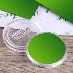 Puede incluir: Dos insignias redondas de plástico transparente con un imperdible. Una insignia tiene un inserto circular verde brillante. La otra insignia está desmontada, mostrando el imperdible y la carcasa de plástico transparente. El fondo incluye papel verde.