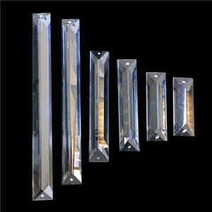 May include: A collection of six clear crystal prism pendants of different lengths, set against a black backdrop. Each rectangular prism has a pointed base and a small hole at the top. The prisms are designed to reflect light, creating a shimmering effect.