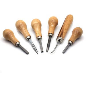 May include: A collection of six wood-handled carving tools. The tools feature different blade shapes, including straight, angled, and curved, made of silver-coloured metal. The handles are light brown, and the tools are displayed against a white backdrop.