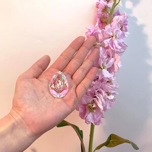 May include: A clear, iridescent teardrop-shaped pendant with an oval design in the center. The pendant is held in a hand, with a backdrop of pink flowers. The pendant reflects multiple colors, including pink, purple, and yellow.