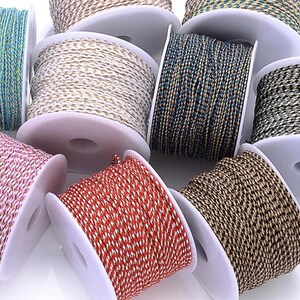 May include: Assortment of spools of decorative cord in various colors, including gold, red, pink, blue, and brown. The cord is tightly wound on white plastic spools, showcasing a variety of textures and patterns. Ideal for crafting and embellishment projects.