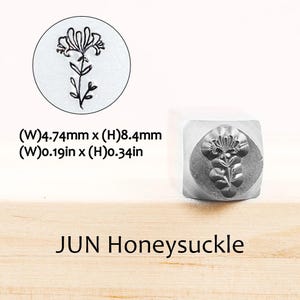 Pode incluir: Um carimbo de metal com um desenho de madressilva. O carimbo tem uma pega quadrada e uma impress&atilde;o circular de uma flor de madressilva. O texto "JUN Honeysuckle" est&aacute; abaixo. O carimbo tem 4,74 mm de largura e 8,4 mm de altura.