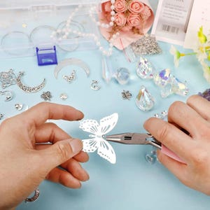 May include: A craft project in progress, featuring a white butterfly cutout being worked on with pliers. The scene includes various jewelry-making supplies: silver charms, crystals, beads, and a small bouquet of pink roses, all arranged on a light blue surface.