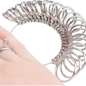 May include: A silver-coloured metal ring sizer set consisting of multiple circular bands of varying diameters attached to a flexible wire cable, with one individual ring band currently placed on a finger for size testing purposes.