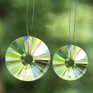 Puede incluir: Dos atrapasoles de cristal iridiscente colgando sobre un fondo verde borroso. Los cristales circulares tienen un diseño radial con un agujero central, reflejando un espectro de colores que incluye verde, amarillo y azul. Están suspendidos por finos hilos transparentes.