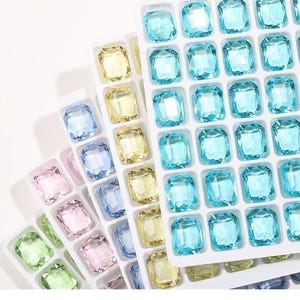 May include: A selection of colourful, faceted gemstones in shades of blue, yellow, pink, and green, arranged in white trays. The gemstones are rectangular with multiple facets, creating a sparkling effect. The trays are stacked, showcasing the variety of colours.