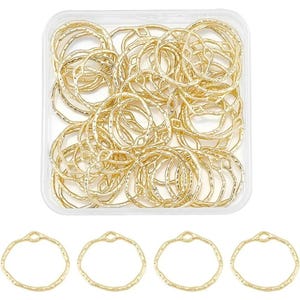 May include: A collection of gold-colored, open-circle jewelry findings in a clear plastic container. The findings have a textured surface and a small, decorative opening at the top. Four additional findings are displayed below the container.