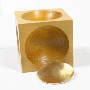 Peut inclure: Un cube en bois de couleur claire présentant une grande indentation circulaire sur la face avant et une indentation circulaire plus petite sur la face supérieure. Un disque métallique doré séparé repose sur la surface devant le cube en bois.