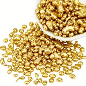 May include: A collection of small, irregular, metallic gold-colored nuggets scattered across a surface, with a larger cluster contained within a white bowl. The pieces have a smooth, reflective surface and vary in size and shape, resembling raw metal.